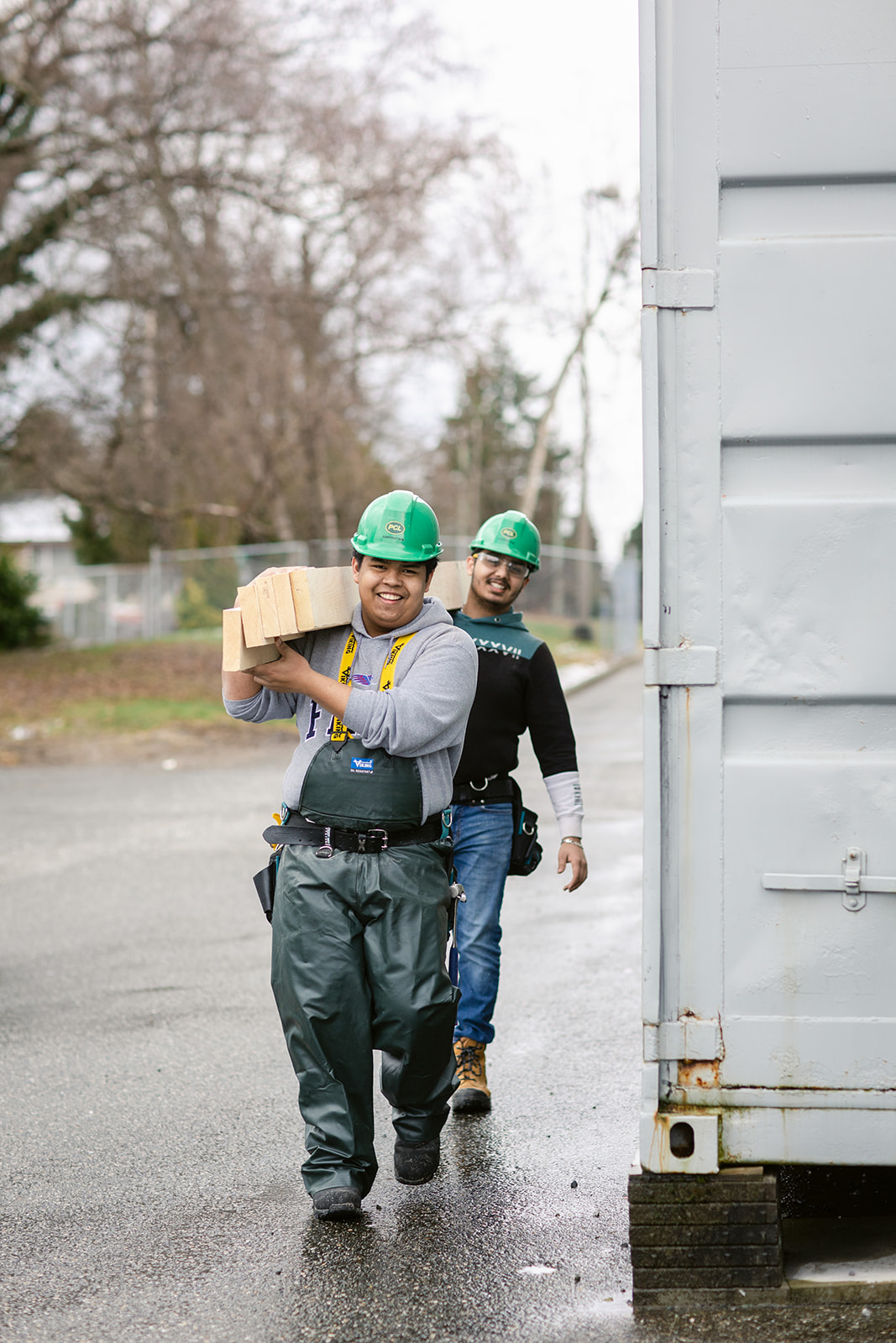 Two young carpenters walking about carrying planks of wood.
