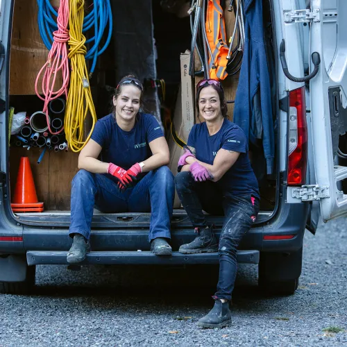 Two female tradespeople sitting at the back of a truck with supplies.